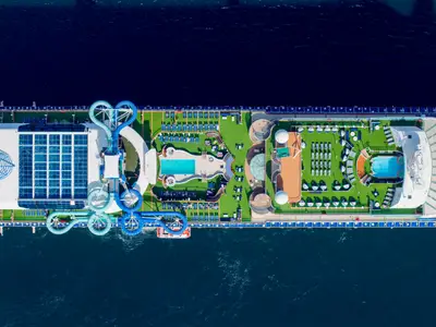 Vogelperspektive auf das Oberdeck der Carnival Adventure. Man sieht farbige Wasserrutschen, Schwimmbecken und Liegeflächen mit vielen Sonnenliegen. Das Schiff ist von blauem Meer umgeben.