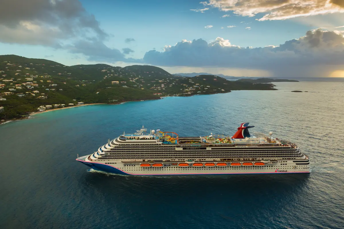 Luftbild der Carnival Breeze auf blauem Meer. Links grüne Hügel mit Häusern, rechts warmer Sonnenuntergangshimmel. Es fährt nach rechts.