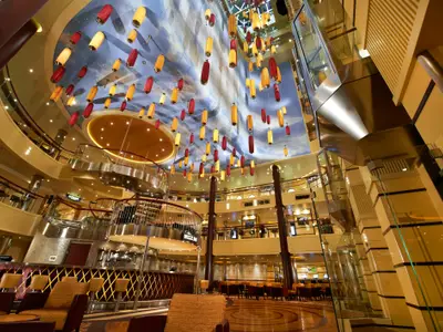 Weitwinkelaufnahme des mehrgeschossigen Atriums der Carnival Breeze. Unter einer Himmelsdecke hängen rote und gelbe Laternen. Unten sind Bars, Sitzbereiche und ein geschwungenes Treppenhaus sichtbar. Glasschächte und Aufzüge sind rechts.