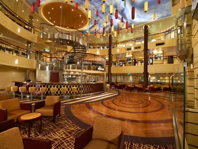 Geräumiges, modernes Atrium auf der Carnival Breeze mit einer runden Bar, bequemen Sitzbereichen und mehreren Etagen mit Balkonen. Eine große Lichtinstallation hängt von der Decke, die wie ein Himmel mit Wolken bemalt ist. Der Boden im Vordergrund ist aus Holz mit konzentrischen Kreismustern.