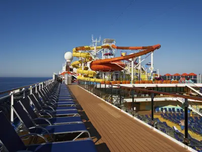 Weitwinkelansicht des sonnigen Sonnendecks der Carnival Breeze mit farbenfrohen Wasserrutschen, zahlreichen blauen Liegestühlen und dem tiefblauen Ozean am Horizont.