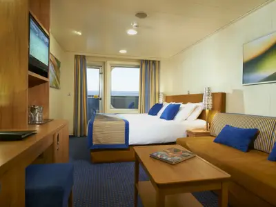 Ansicht einer modernen Balkonkabine auf der Carnival Breeze. Die Kabine verfügt über ein Doppelbett, ein Sofa, einen Schreibtisch und Zugang zu einem Balkon mit Meerblick. Blaue Akzente dominieren das Design.