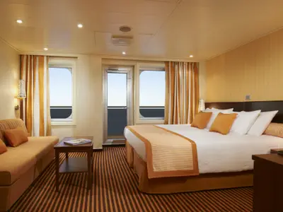 Eine moderne Suite auf der Carnival Breeze bei Tageslicht. Zu sehen sind ein Doppelbett, ein Sofa, eine Tür zum Balkon und Fenster mit Meerblick.