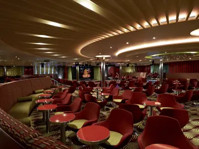 Weitwinkelaufnahme einer lebhaften Bar-Lounge auf der Carnival Breeze mit einer beleuchteten Bühne im Hintergrund, umgeben von Reihen roter Sessel und kleiner Tische. Stimmungsvolle Beleuchtung und gemusterter Teppich.