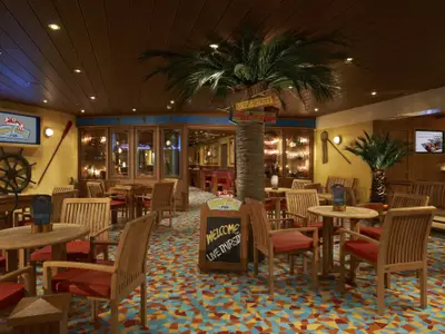 Innenansicht der tropischen 'Red Frog Pub' Bar auf der Carnival Breeze. Holztische, Stühle mit roten Polstern, große Palmen, bunter Fliesenboden und maritime Wanddekoration prägen den Raum.