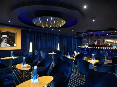 Eine stilvolle, dunkelblaue Bar-Lounge auf der Carnival Breeze mit Samtsesseln, runden Tischen, funkelnden Vorhängen und einem gerahmten Bild von Elton John an der Wand. Ein runder Tresen mit Barhockern ist rechts sichtbar.