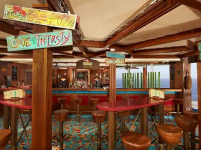 Innenansicht der RedFrog Rum Bar auf der Carnival Breeze mit bunter Einrichtung, Holzdekor und Meerblick durch große Fenster. Mehrere leere Barhocker und Tische sind zu sehen.
