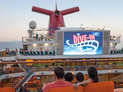 Familie schaut DIVE-IN Movies auf großer Leinwand auf einem Deck der Carnival Breeze bei Dämmerung. Ihr roter Schornstein ist im Hintergrund zu sehen.