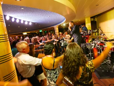 Ein Sänger tritt auf einer beleuchteten Bühne auf der Carnival Breeze auf, während eine Gruppe von Menschen davor tanzt und die Arme in die Luft streckt. Im Hintergrund ist ein Schlagzeug zu sehen.