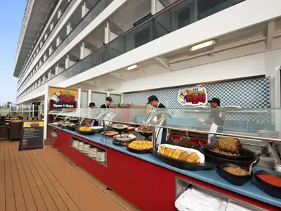 Sonnendeck der Carnival Breeze mit Fat Jimmy's BBQ-Buffet. Crewmitglieder servieren Fleisch, Beilagen. Ihre Seitenwand und Sitzplätze im Hintergrund.