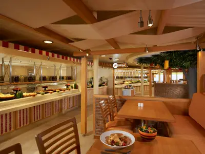 Helles Buffetrestaurant auf der Carnival Breeze mit langen Theken voller Speisen in Gläsern und Schüsseln. Sitzbereiche und ein künstlicher Baum.