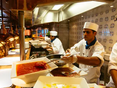 Zwei Köche in weißen Uniformen und Mützen servieren Speisen an einem Buffet. Im Vordergrund sind Töpfe mit roter Sauce und Fleischgerichten unter goldenen Wärmelampen zu sehen. Ein Koch in Handschuhen füllt eine Schale.