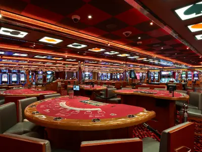 Großes, leeres Casino der Carnival Breeze. Rote Spieltische im Vordergrund, viele Slotmaschinen im Hintergrund. Decke mit rot-schwarzem Schachbrettmuster und leuchtenden Kartensymbolen. Überwachungskameras sichtbar.