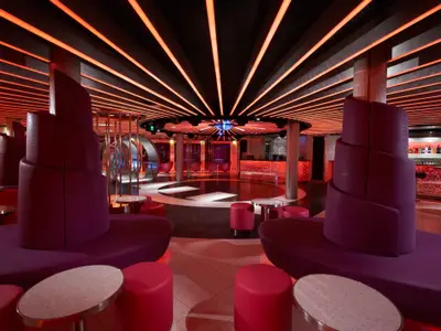 Ein moderner Nachtclub auf der Carnival Breeze mit leuchtend roter Beleuchtung, einer runden Tanzfläche und bequemen, geschwungenen Sitzgelegenheiten in Violett- und Rottönen.