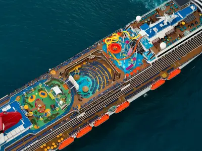Luftaufnahme des lebhaften Oberdecks der Carnival Breeze mit gelben und roten Wasserrutschen, einem Minigolfplatz und vielen blauen Liegestühlen am Pool. Das Meer umgibt das Schiff.