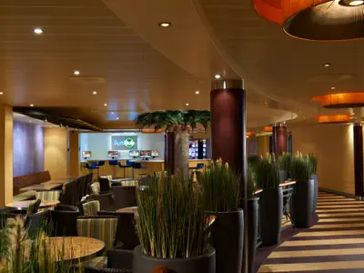 Eine helle, moderne Lounge auf der Carnival Breeze mit verschiedenen Sitzbereichen, runden Tischen und hohen Pflanzen in Töpfen. Im Hintergrund ist eine Theke mit Computern und dem Schriftzug 'Fun Hub' zu sehen.