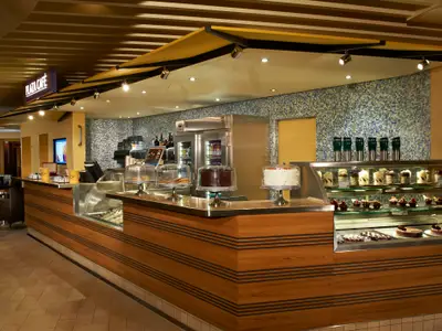Modernes Café auf der Carnival Breeze. Eine Holztheke präsentiert Kuchen und Gebäck in Vitrinen. Kaffeemaschine und Mosaikwand im Hintergrund. Warme, einladende Beleuchtung.