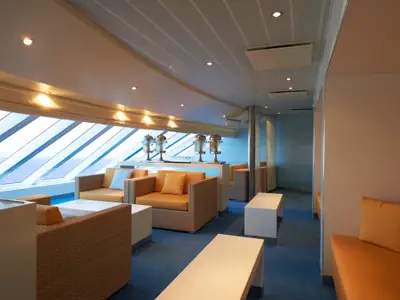 Geräumige, modern eingerichtete Lounge auf der Carnival Breeze mit großen Schrägfenstern mit Blick auf das offene Meer. Mehrere orangefarbene Sitzgruppen und weiße Tische sind auf blauem Teppichboden verteilt. An einer Theke stehen Getränkespender.