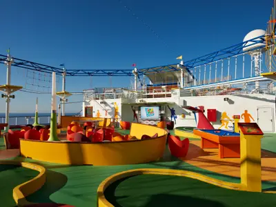 Blick auf das bunte Oberdeck der Carnival Breeze mit einer Minigolf-Anlage, Sitzgelegenheiten, einem Seilgarten und einem großen Bildschirm. Im Hintergrund ist das blaue Meer unter klarem Himmel sichtbar.