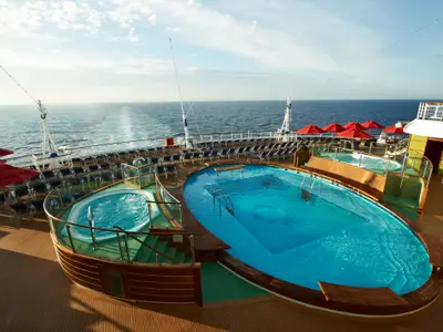 Großer Poolbereich mit zwei Whirlpools auf dem Sonnendeck der Carnival Breeze. Viele Liegestühle und rote Sonnenschirme. Das Schiff fährt auf dem offenen Meer.