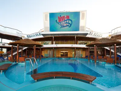 Großer Poolbereich auf der Carnival Breeze mit einem riesigen LED-Bildschirm, der "Dive-In Movies" zeigt. Strohgedeckte Bars, Liegestühle und Sitzbänke im Wasser umgeben den Pool. Mehrere Decks sind sichtbar.