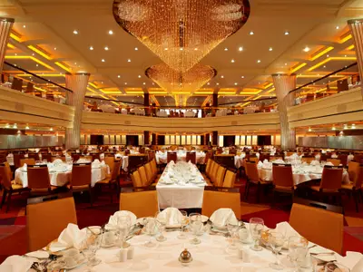 Geräumiges, elegantes Hauptrestaurant auf der Carnival Breeze. Mehrere Ebenen mit weiß gedeckten Tischen und orangen Stühlen sind sichtbar. Zwei große, goldene Kronleuchter hängen von der Decke. Keine Personen anwesend.