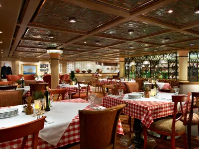 Innenansicht eines traditionell eingerichteten Restaurants. Zahlreiche Tische sind mit rot-weiß karierten Tischdecken und weißen Deckservietten gedeckt. Dazu stehen Holzstühle bereit. Im Hintergrund sind maritime Dekorationen, darunter ein Gemälde eines Segelschiffs und eine Wand mit Weinregalen, sowie eine kunstvoll verzierte Decke zu sehen. Keine Personen anwesend.