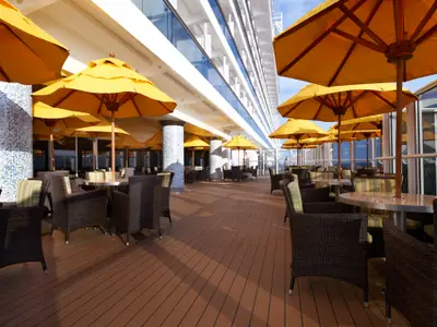 Eine weitläufige Außenlounge auf der Carnival Breeze bei Tag. Zahlreiche braune Rattansessel und Tische mit gelben Sonnenschirmen stehen auf einem Holzdeck. Im Hintergrund ein modernes Schiffsgebäude mit Glasfronten und Meerblick.