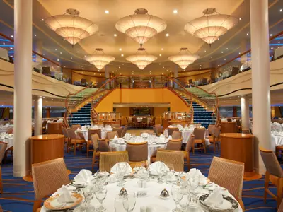 Ansicht eines eleganten, mehrstufigen Restaurants auf der Carnival Breeze. Mehrere Ebenen mit weiß gedeckten Tischen, blauer Teppich, eine imposante goldene Doppeltreppe und große Deckenlampen sind zu sehen. Das Ambiente ist gehoben.