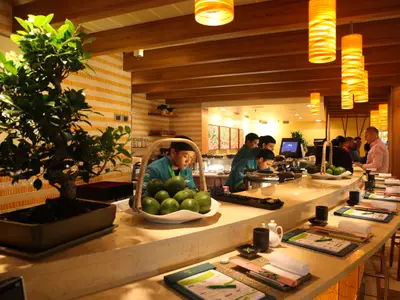 Eine helle, moderne Sushi-Bar in einem Restaurant der Carnival Breeze. Mehrere Crew-Mitglieder bereiten Speisen an einem langen Tresen zu. Ein Korb mit Avocados und ein Bonsai-Baum sind auf dem Tresen zu sehen. Orangefarbene Lampen hängen von der Decke.