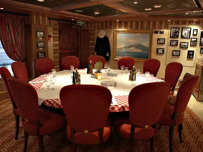 Ein festlich gedeckter Rundtisch mit roten Stühlen in einem Restaurant an Bord. Die Wände sind mit maritimen Bildern und einer Uniform dekoriert. Ein großes Fenster gibt den Blick auf das offene Meer frei.