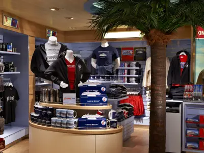 Ein heller Shop auf der Carnival Breeze zeigt Kleidung, Tassen und andere Souvenirs der 'Carnival Breeze'. Mehrere Mannequins präsentieren Hoodies und T-Shirts. Eine große Palmenattrappe steht mittig. Die Regale sind gefüllt mit Fanartikeln.