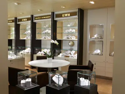 Blick in einen hell beleuchteten Schmuckladen auf der Carnival Breeze. Mehrere Vitrinen zeigen Halsketten, Ringe und andere Schmuckstücke. Ein runder Tisch mit braunen Sesseln und einer weißen Orchidee steht im Zentrum. An der Wand sind weitere Vitrinen und Schränke zu sehen.