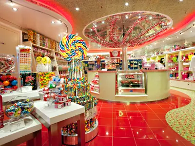 Ein bunter Süßigkeitenladen an Bord der Carnival Breeze. Im Vordergrund ein riesiger Spiral-Lutscher, umgeben von Regalen voller Bonbons, Schokolade und anderen Leckereien. Der Boden ist teils rot gefliest, teils mit einem gemusterten Teppich ausgelegt. Eine gläserne Deckenkonstruktion mit roten Verzierungen spiegelt das Licht.