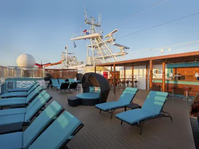 Blick auf das sonnige Oberdeck der Carnival Breeze mit Reihen von blauen Liegestühlen, runden Loungesesseln und einer Bar. Im Hintergrund sind der Schiffsmast und eine Wasserrutsche sichtbar.