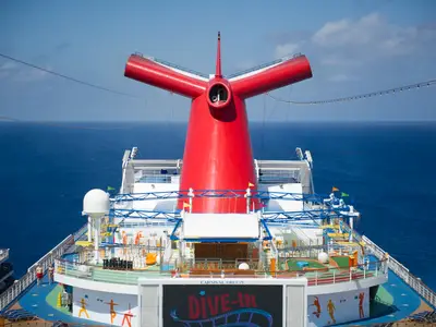 Weitblick auf das Heck der Carnival Breeze bei strahlendem Sonnenschein. Dominant ist der rote, Y-förmige Schornstein. Darunter befinden sich Freiluftdecks mit Minigolf und einem großen Bildschirm. Im Hintergrund das tiefblaue Meer.