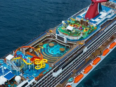 Luftaufnahme des Oberdecks der großen Carnival Breeze mit farbenfrohen Wasserrutschen, einem weitläufigen Poolbereich mit Liegestühlen und einer Minigolfanlage bei Tageslicht. Der Ozean umgibt das Schiff.