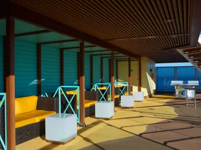 Überdachtes Sonnendeck auf der Carnival Breeze mit türkisen Wänden, braunen Holzpfeilern, gelben Outdoor-Sitzkissen und dunklen Rattanmöbeln. Rechts ein Pingpong-Tisch und Blick auf das blaue Meer und den Himmel.