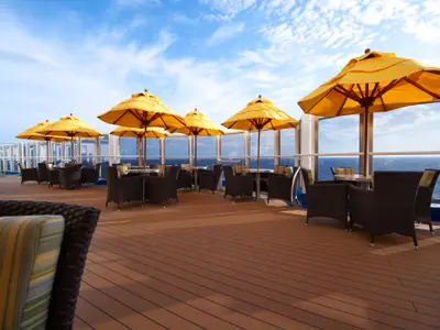 Ansicht eines Sonnendecks auf der Carnival Breeze. Mehrere Tische und Rattan-Sessel mit gestreiften Kissen stehen unter gelben Sonnenschirmen. Im Hintergrund das weite Meer und blauer Himmel mit Wolken, durch eine Glasreling sichtbar.