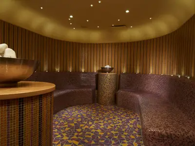 Runder Spa-Raum mit Mosaikfliesen, geschwungenen Sitzbänken und warmem Licht. Eine große Schale mit weißen Steinen steht auf einem Holztisch.