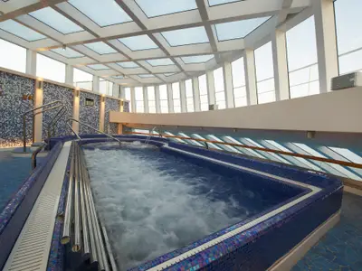 Ein geräumiger Wellnessbereich auf der Carnival Breeze mit einem großen, sprudelnden Whirlpool im Vordergrund. Blaue Mosaikfliesen bedecken den Poolrand und die Wände. Die Decke besteht aus Glas, und hohe Fenster bieten Weitblick. Edelstahl-Haltegriffe führen ins Wasser.