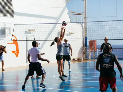 Mehrere Erwachsene spielen Volleyball auf einem blauen Sportfeld der Carnival Breeze. Eine Frau springt, um den Ball zu schlagen, während andere Spieler zusehen. Im Hintergrund sind das Meer und der Himmel zu sehen.