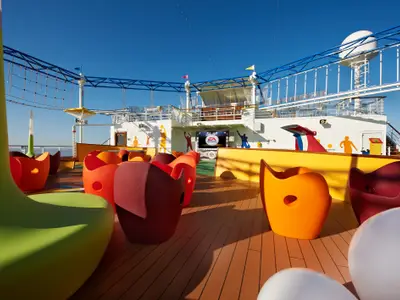 Ein lebhaftes Outdoor-Deck der Carnival Breeze am Tag. Bunte, moderne Sitzgelegenheiten aus Kunststoff dominieren den Vordergrund. Im Hintergrund eine große Leinwand mit dem 'EA Sports'-Logo, umgeben von sportlichen Silhouetten an der weißen Wand des Schiffes. Ein Volleyballnetz ist ebenfalls sichtbar unter blauem Himmel.