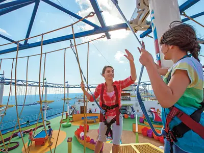 Zwei Frauen in Sicherungsgurten balancieren auf einem Hochseilgarten auf dem oberen Deck der Carnival Breeze. Im Hintergrund ist das Meer und eine Minigolfanlage sichtbar. Eine Frau winkt der anderen zu.