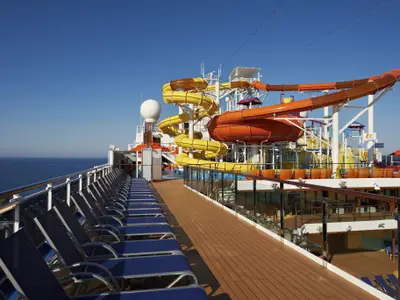 View of a lively cruise ship deck featuring vibrant yellow and orange water slides, a wide sun deck with lounge chairs, and the expansive blue ocean under a clear sky.