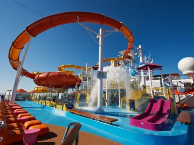 Ein bunter Wasserpark auf dem Oberdeck der Carnival Breeze mit orangefarbenen und gelben Wasserrutschen, Wasserspielen und einem lila Becken unter blauem Himmel.