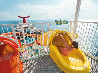 Eine Frau in rotem Badeanzug rutscht eine gelbe Wasserrutsche auf der Carnival Breeze. Man sieht weitere Rutschen, Pools und das blaue Meer. Die Szene vermittelt lebhaften Urlaubsspaß.