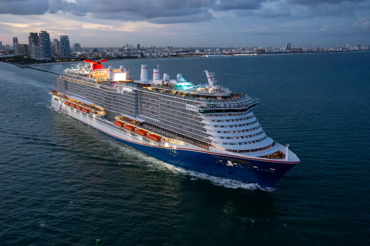 Luftaufnahme der Carnival Celebration, die bei Dämmerung auf See vor der beleuchteten Skyline einer Küstenstadt fährt.