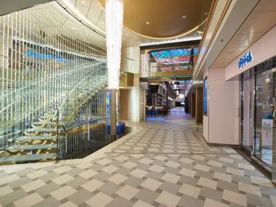 Helles, modernes Atrium der Carnival Celebration. Links gläserne Treppe, mittig hoher Kristallleuchter. Gemusterter Boden fuehrt zu Bar und Laden unter farbigem Deckendekor. Ohne Personen.