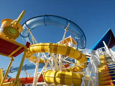 Bunter Aqua Park auf der Carnival Celebration. Große Wasserrutschen in Blau und Gelb. Riesiger gelber Kippeimer links. Bunte Treppen führen hoch. Strahlend blauer Himmel mit kleinem Mond.